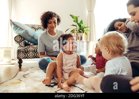 Mütter im Wohnzimmer, die ihre spielenden Babys betrachten. Freundinnen treffen sich am Wochenende mit ihren Kindern. Stockfoto