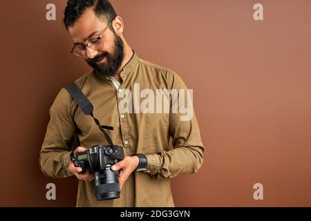 Fotograf mit Kamera isoliert auf braunem Hintergrund, erfolgreiche professionelle männliche Blicke auf Fotos auf dem Bildschirm der Kamera. Fotografie Konzept Stockfoto