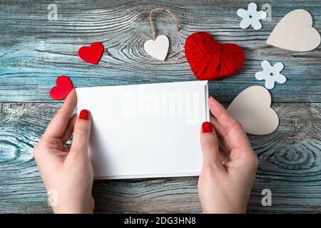 Weißer Umschlag mit einem leeren Brief in den Händen einer Frau auf einem romantischen Hintergrund mit Herzen und einem Band. Stockfoto