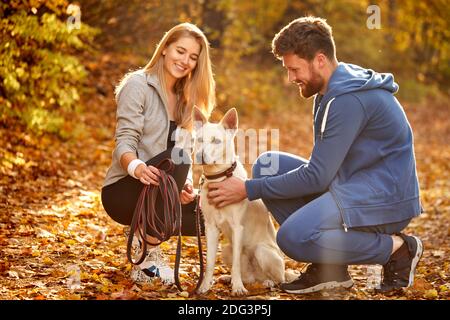 Freundliches nettes Paar mit weißem Haustier Hund in der Natur, Herbst sonnigen Tag, Spiel und Schlaganfall. Perfektes Wetter für Spaziergänge Stockfoto