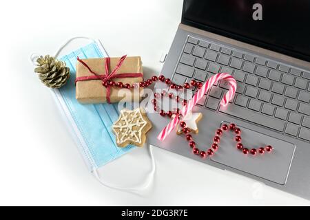 Online-Shopping für Weihnachten während Coronavirus Pandemie, Laptop mit Schutzmaske, Geschenk, Süßigkeiten und Urlaubsdekoration auf einem weißen Backgroun Stockfoto