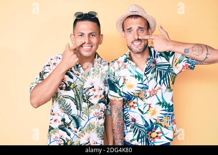 Junge schwule Paar von zwei Männern tragen Sommerhut und hawaiiihemd zeigt mit Hand Finger zu Gesicht und Nase, lächelnd fröhlich. Beauty-Konzept Stockfoto