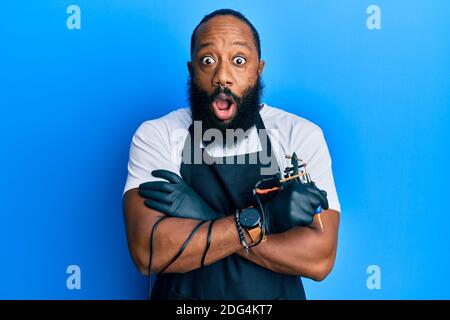 Junge afroamerikanische Mann Tattoo Künstler tragen professionelle Uniform und Handschuhe halten Tätowiermaschine Angst und schockiert mit Überraschung und Begeistert Stockfoto