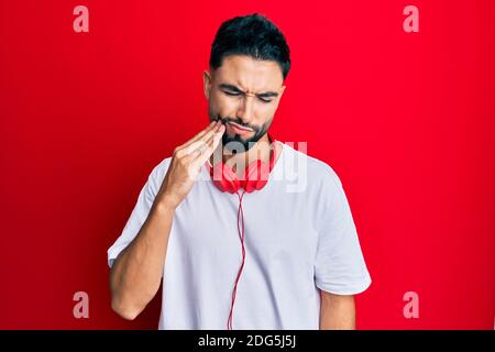 Junger Mann mit Bart hört Musik mit Kopfhörer berühren Mund mit Hand mit schmerzhaftem Ausdruck wegen Zahnschmerzen oder Zahnerkrankung auf te Stockfoto