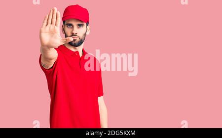 Junge gutaussehende Mann mit Bart tragen Lieferung Uniform tun aufhören zu singen mit Handfläche der Hand. Warnung Ausdruck mit negativen und ernsthaften Geste auf Stockfoto