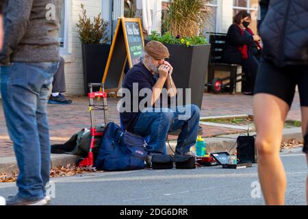 Alexandria, VA, USA 11-28-2020: Ein weißer bärtiger alter Straßenmusiker mit Stiefeln, Jeans und flachem Hut sitzt auf dem Hocker am Bürgersteig spielt Stockfoto