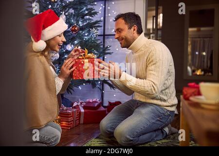 Hübscher Mann, der Geschenk für schöne Frau gibt, während er innen sitzt Dekoration Weihnachtszimmer Stockfoto