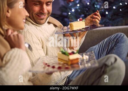 Glückliches lächelndes Paar, das Kuchen zusammen im Wohnzimmer auf isst Weihnachtsbaum Hintergrund Stockfoto