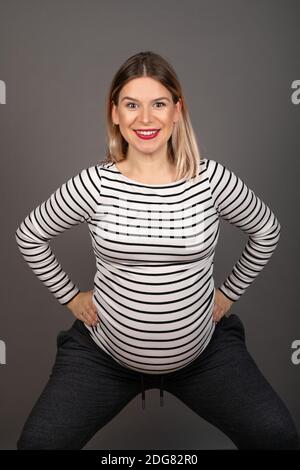 Schwangere Frau, die sich auf die Arbeit vorbereitet, hockt vor grauem Hintergrund Stockfoto