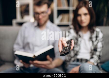 Bärtiger Mann, der neues Buch liest, während er auf der Couch in der Nähe der schönen Frau sitzt, die Fernsehen schaut. Freizeit zu Hause. Junges Paar zusammen entspannen. Stockfoto