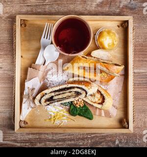 Mohn- und Apfelstrudel auf einem rustikalen Holztablett. Tee in einer Papiertasse mit einem weißen leeren Platz für Text und einem Löffel und einer Gabel aus umweltfreundlichen Materialien. Stockfoto