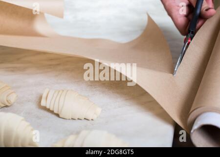 Schere Schneiden Sie ein Backpapier und rohes Croissant auf einem weißen Tisch. Stockfoto