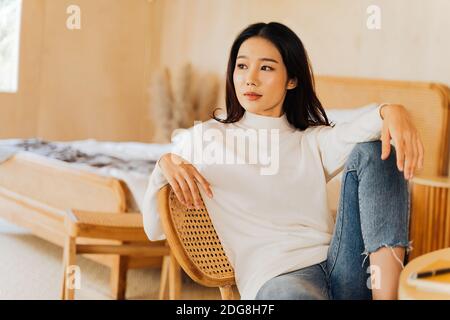 20s junge Teenager asiatische Frau trägt einen Pullover Blick weg in stilvollen gemütlichen Raum Hintergrund. Attraktive weibliche Modell posiert für Kamera auf Stuhl. Winter Portrait Mode Lifestyle. Platz für die Banner-Kopie Stockfoto