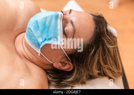 Eine Frau liegt mit Akupunkturnadeln auf einer Bahre im Zimmer eines Physiotherapeuten und trägt eine Coronavirus-Schutzmaske Stockfoto