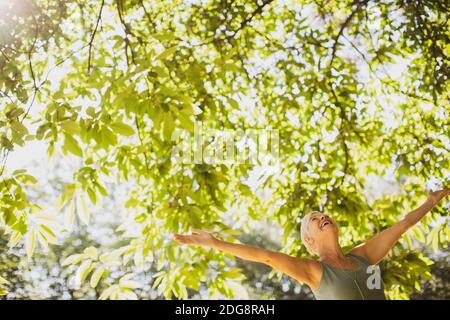 Überschwängliche ältere Frau mit ausgestreckten Armen unter einem sonnigen Baum Stockfoto