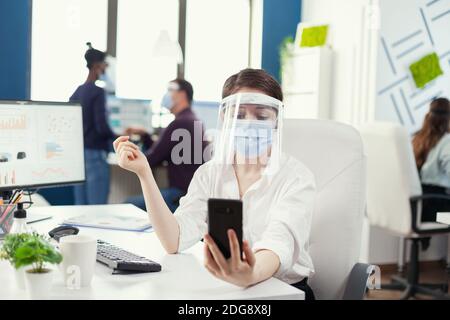 Geschäftsfrau mit einer Unternehmensvideokonferenz mit Smartphone trägt Gesichtsmaske. Kollegen, die während einer globalen Pandemie mit Coronavirus soziale Distanz respektieren. Stockfoto