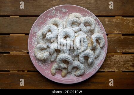 Ein hellrosa Teller Vanillekipferl (kleine, halbmondförmige Weihnachtsbonbons aus gemahlenen Nüssen und Vanille) auf Holzhintergrund. Stockfoto