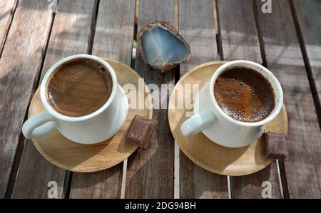 Schäumender türkischer Kaffee aus frisch gerösteten und fein gemahlenen Kaffeebohnen, serviert in kleinen Keramikbechern auf Bambusplatten mit Bitterschokolade-Stücken Stockfoto