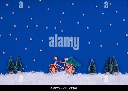 Lustige gemütliche Szene mit Weihnachtsspielzeug auf klassischem Blau. Weihnachtsbaum wird mit dem Fahrrad auf der Schneestraße mit Sternen nach Hause bewegt. Horizontal Stockfoto