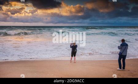 Ein Panoramabild eines Urlauters, der mit seinem Handy einen spektakulären Sonnenuntergang über Fistral Beach in Newquay in Cornwall fotografiert. Stockfoto