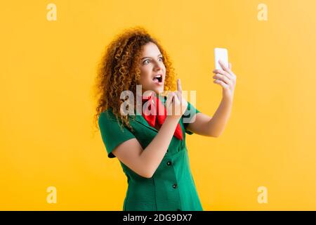 Unhöfliche junge Frau zeigt Mittelfinger während der Aufnahme Selfie auf Gelb Stockfoto