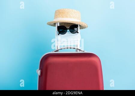 Sonnenbrille, medizinische Maske und Strohhut auf rotem Gepäck auf Blau Stockfoto