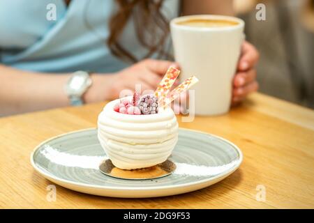 Süßer weißer Cupcake mit Beeren auf dem Teller am Tisch auf dem Hintergrund einer Frau mit einer Tasse Kaffee In einem Café Stockfoto