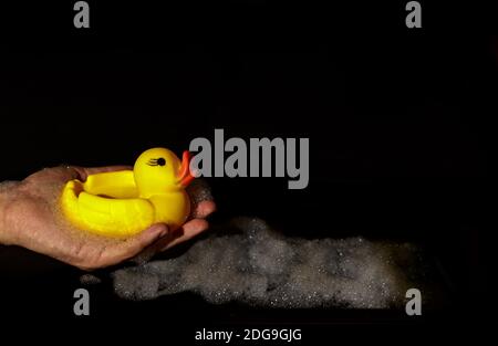 Die Hand des Mannes hält eine gelbe Gummiente auf einer schwarzen Hintergrund mit Schaumstoff Stockfoto