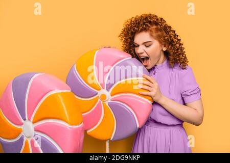 Lollipop, ich will dich essen. Lockige rothaarige positive Frau trägt ein violettes Kleid mit süßen großen Lutscher auf orangefarbenem Hintergrund Stockfoto