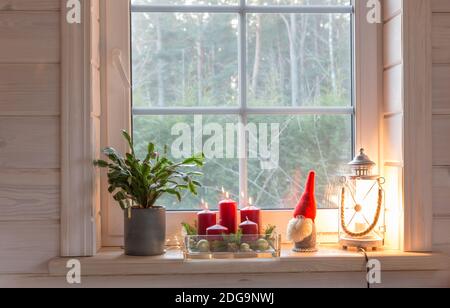 Weihnachtslaterne, Engel, Weihnachtsgnome, Weihnachtskaktus und roter Becher am Fenster eines Holzhauses mit Blick auf den Wintergarten. Stockfoto