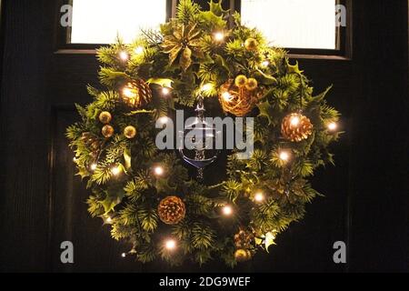 Künstliches Licht-up noblen Weihnachtskranz mit Goldblumen und Pinienzapfen hing an einer schwarzen Tür mit Klopfer in der Mitte in Manchester, England Stockfoto