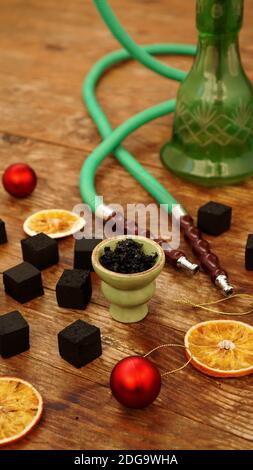 Shisha Hintergrund für Neujahr und weihnachten. Shisha, Holzkohle, trockene Orangenscheiben und weihnachtliche rote Kugeln auf einem Holzhintergrund. Vertikales Foto Stockfoto