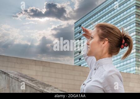Zielgerichtete Frau auf städtischem Hintergrund und Blick nach vorne auf die Sonne zwischen Wolken am Himmel. Geschäftsfrau im Stadtbild. Erfolgreiches Frauenkonzept. Stockfoto