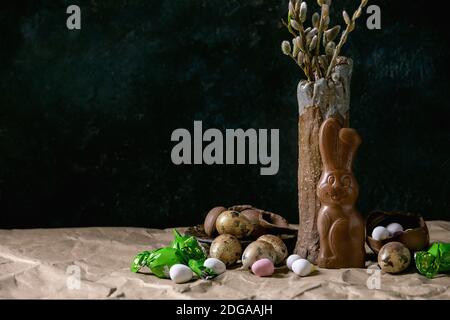 Osterstimmung Stillleben mit Blütenweidenzweigen in Keramikvase, traditionellem Schokoladenkaninchen, Eiern und Süßigkeiten auf dem Tisch mit zerknittertem Bastelpapier. Stockfoto