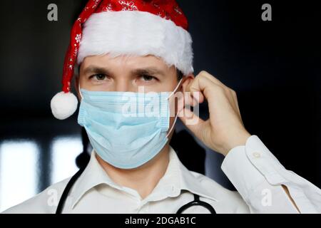 Porträt eines Arztes in medizinischer Gesichtsmaske und Weihnachtsmannhut. Weihnachtsfeier in der Klinik während Coronavirus Pandemie Stockfoto