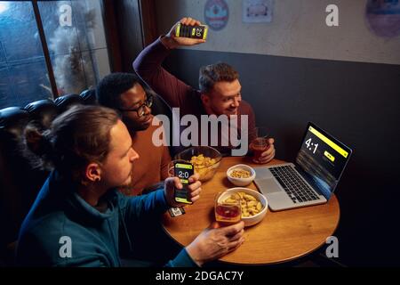 Analysieren. Begeisterte Fans in der Bar mit Bier und mobile App für Wetten, Punkten auf ihren Geräten. Bildschirm mit Match-Ergebnissen, emotionale Freunde jubeln. Glücksspiel, Sport, Finanzen, modernes techn Konzept. Stockfoto