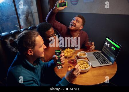 Analysieren. Begeisterte Fans in der Bar mit Bier und mobile App für Wetten, Punkten auf ihren Geräten. Bildschirm mit Match-Ergebnissen, emotionale Freunde jubeln. Glücksspiel, Sport, Finanzen, modernes techn Konzept. Stockfoto