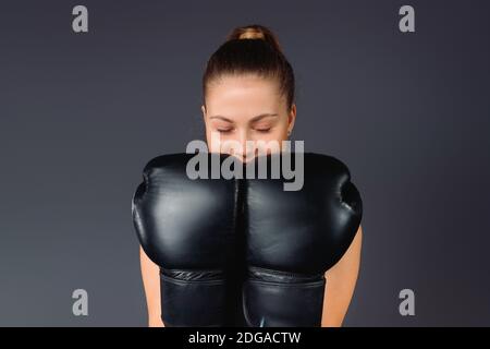 Junge Geschäftsfrau mit geschlossenen Augen, freut sich über ihren Erfolg, hält Boxhandschuhe in der Nähe des Gesichts, auf grauem Hintergrund. Stockfoto