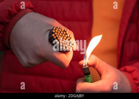 Mann in Rot aufgebockt Beleuchtung bis viele kleine schwarze Feuerwerkskörper in seiner Hand mit Gas Feuerzeug. Guy immer bereit für das neue Jahr Spaß mit Feuerwerk oder pyrotechnische Produkte Stockfoto