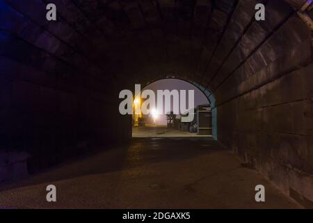 Stimmungsvolle, abendliche Straßenszene mit Blick durch einen alten Backsteintunnel bei Nacht. Blick auf die Straßenlaterne durch einen dunklen Steintunnel. Stockfoto