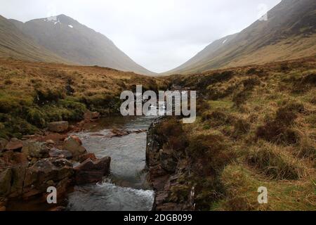 Unter einem weißen Himmel schlängelt sich ein Fluss durch Nebel und flaches Licht im Tal von Glen Coe in der Highland Region (Lochaber Geopark, Schottland) Stockfoto