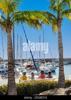 Reife Gruppe Urlaub Living. Spanien Ansicht ältere Rentner Urlaub ältere Urlauber im Ausland Senioren sitzen Winter Sonne Urlaub Gruppe von Reifen Rentner Freunde Urlauber sitzen auf einer sonnigen Bank von Palmen eingerahmt Yachthafen genießen Ansicht Puerto Calero Lanzarote Kanarische Inseln Spanien Konzept zukünftige Finanzierung Renteneinkommen will Lebensstil Stockfoto