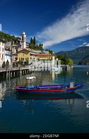 Riva di Solto, eine typisch italienische Kleinstadt am Iseosee mit einer Kirche und einer Strandpromenade. Ein blaues und rotes Boot vor dem Hotel. Bergamo, Lombardei, Italien. Stockfoto