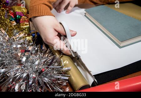 Junges Mädchen schneidet goldenes Geschenkpapier mit weißer Schere Auf einem Tisch mit schwarzem Hintergrund Stockfoto