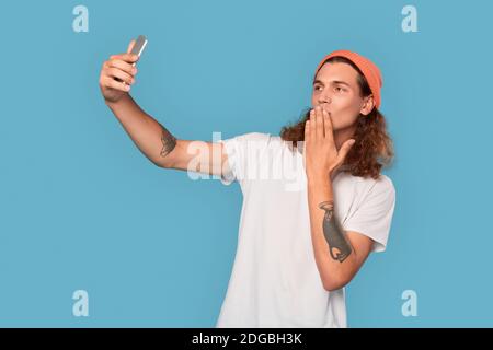 Junge lockige Kerl trägt Hut isoliert auf blauem Hintergrund stehen Nehmen Selfie Foto auf Smartphone küssen Hand posiert Kamera Verspielt Stockfoto