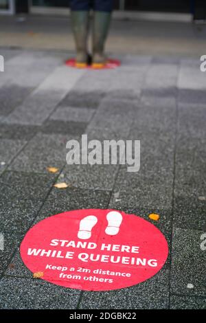 Soziale Wegmarkierungen auf dem Boden im Stadtzentrum von Großbritannien. Steht hier, während ihr ansteht. Halten Sie einen Abstand von 2 m zu anderen Personen ein. Stockfoto