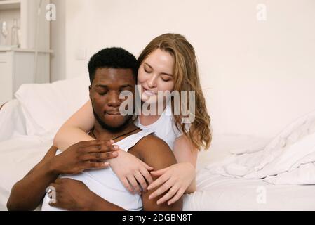 Porträt eines gemischten Rennpaares im Wohnzimmer sitzend Umarmen Stockfoto