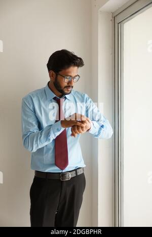 Portrait of Indian Bengali großer, dunkler, hübscher Brünette junger Mann in Bürokleidung, der die Uhr vor dem Glasfenster im Büro sieht. Stockfoto