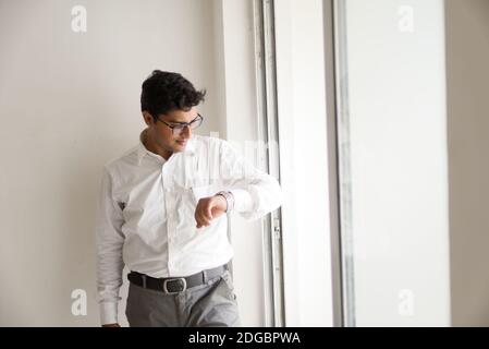Portrait of Indian Bengali großer, dunkler, hübscher Brünette junger Mann in Bürokleidung, der die Uhr vor dem Glasfenster im Büro sieht. Stockfoto