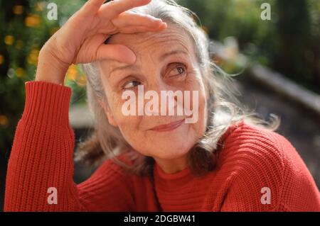 Porträt der älteren Frau mit grauen Haaren und Gesicht mit Falten tragen roten Strickpullover und entspannen im Park während Sonniger Tag Stockfoto